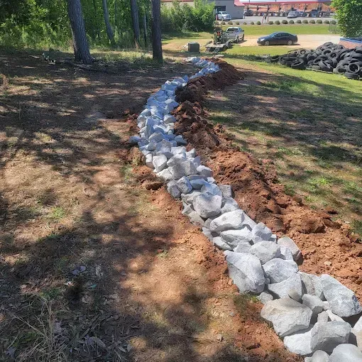 Rock-lined drainage ditch dug in dirt, alongside grass and trees.