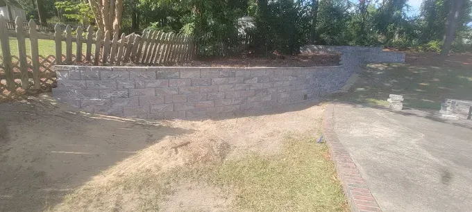 A retaining wall made of gray blocks with a wooden fence and grass in the foreground and trees in the background.