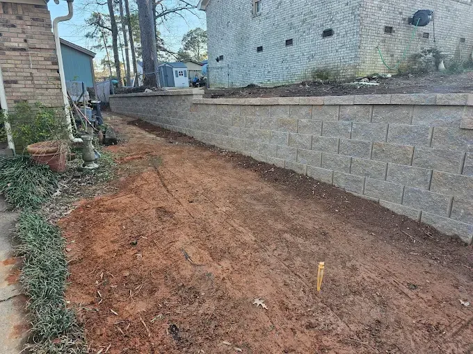 A dirt pathway between a retaining wall and building. Brown soil, gray blocks, and green foliage.