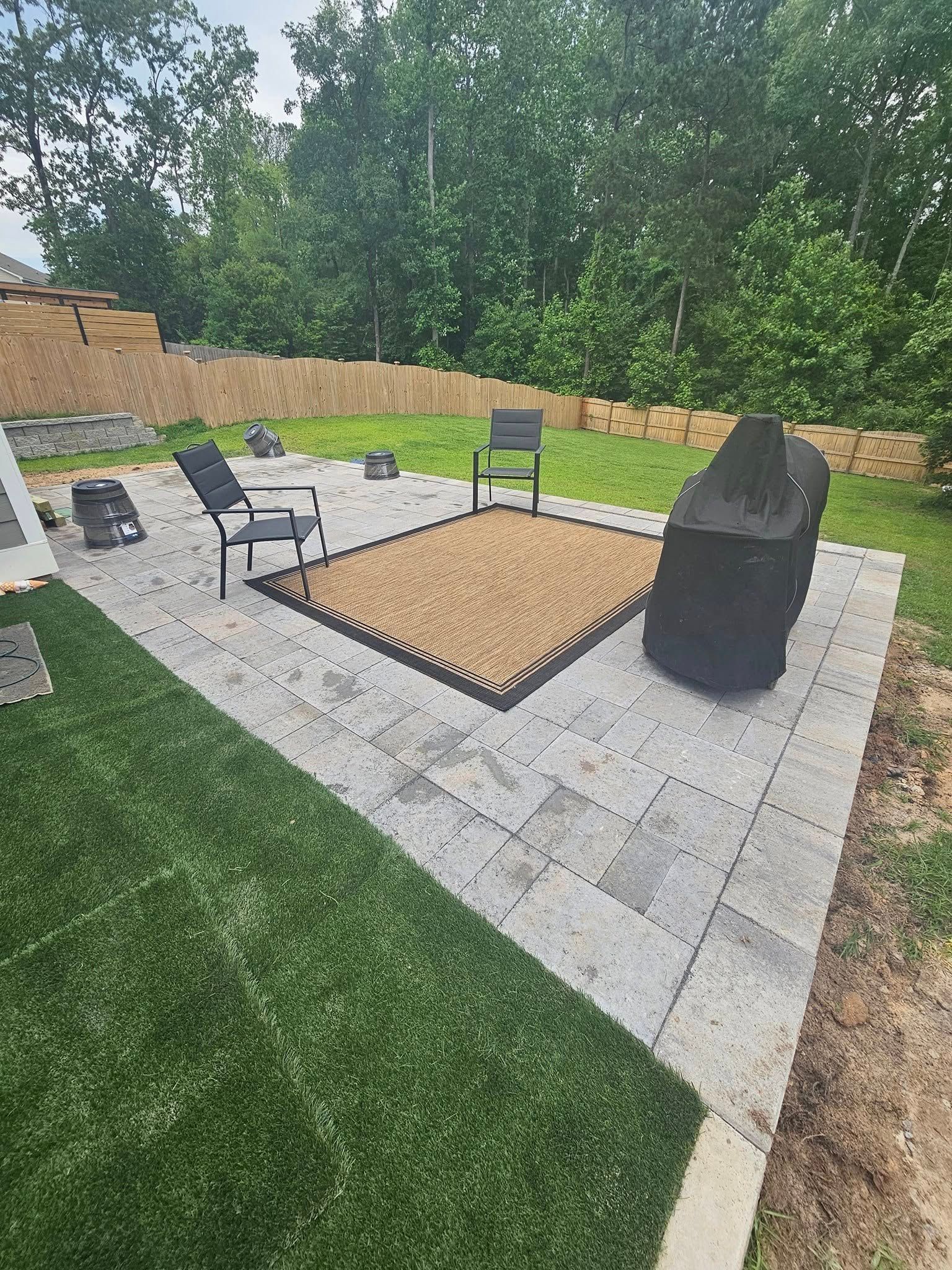 Patio with gray pavers, artificial grass, and outdoor furniture. Black grill and chairs centered on textured mat.