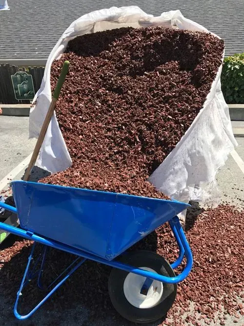 Reddish-brown gravel spilling from a large bag into a blue wheelbarrow, shovel leaning nearby. Outdoors.