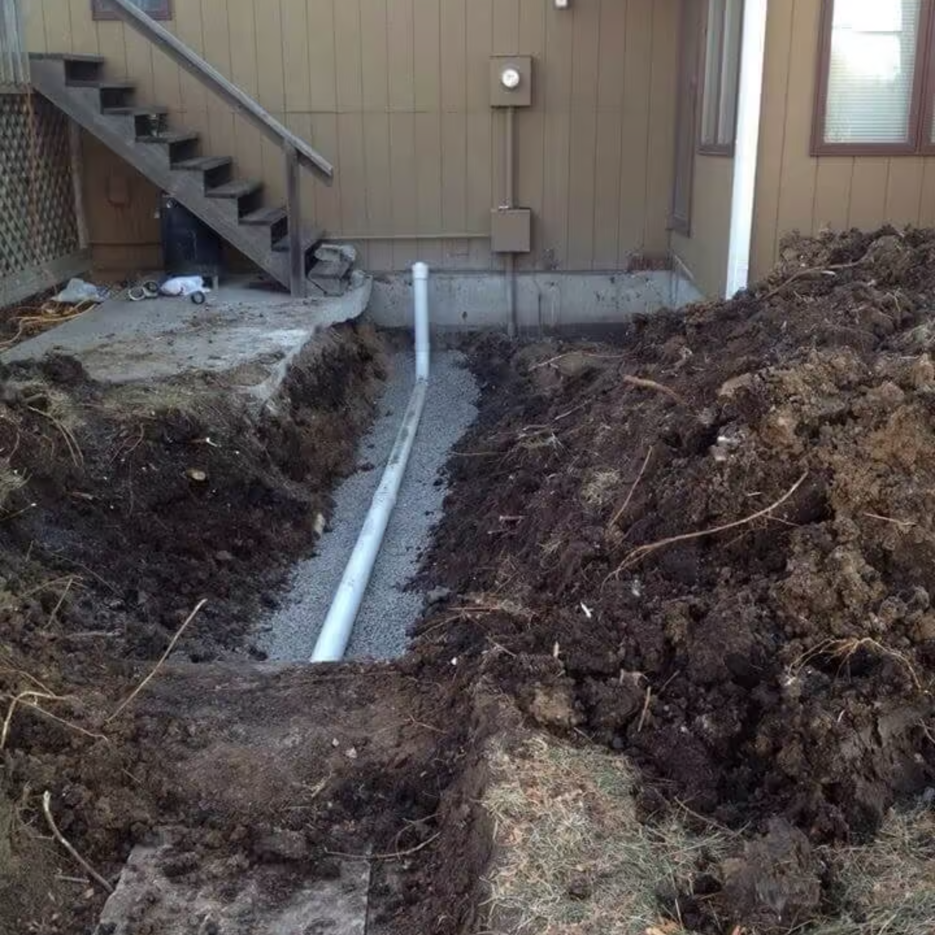 Trench with a white pipe installed, near a house with stairs. Dirt piles surround the trench.