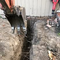 An excavator bucket digs a narrow trench beside a building.