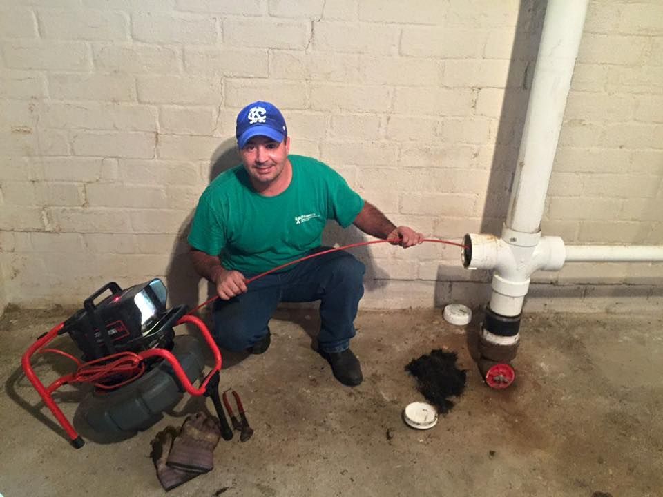 Plumber in green shirt and Clemson hat, cleaning a pipe in a basement.