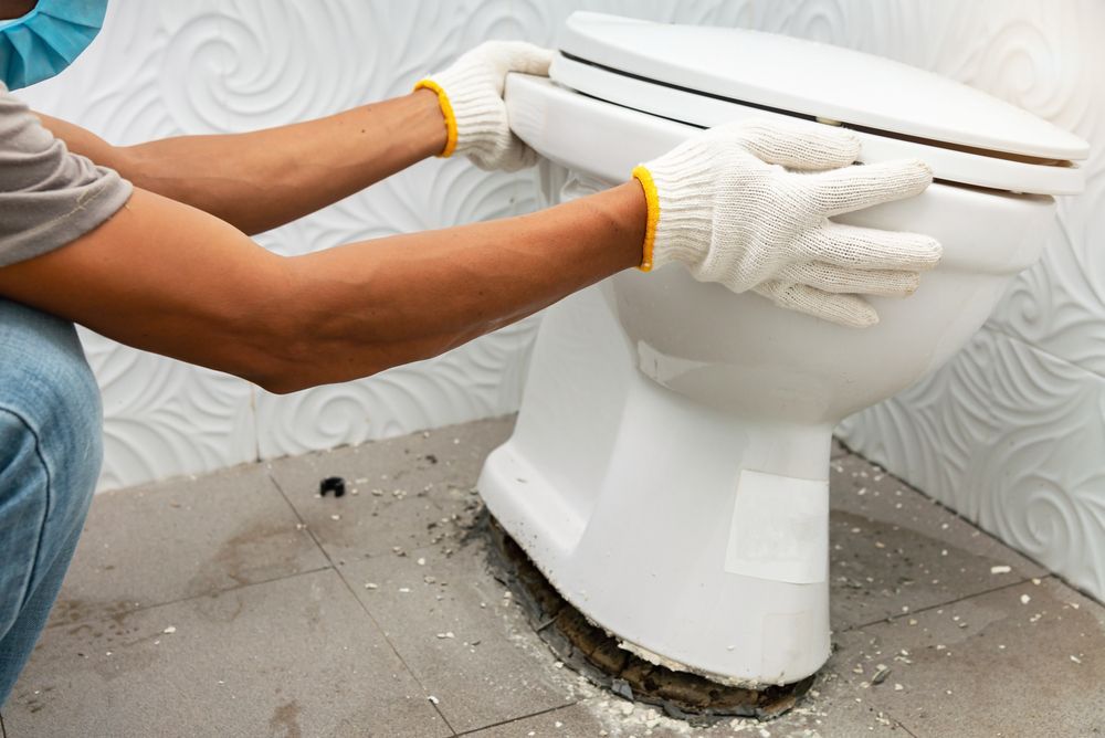 Person wearing gloves installing a white toilet in a bathroom, near debris.