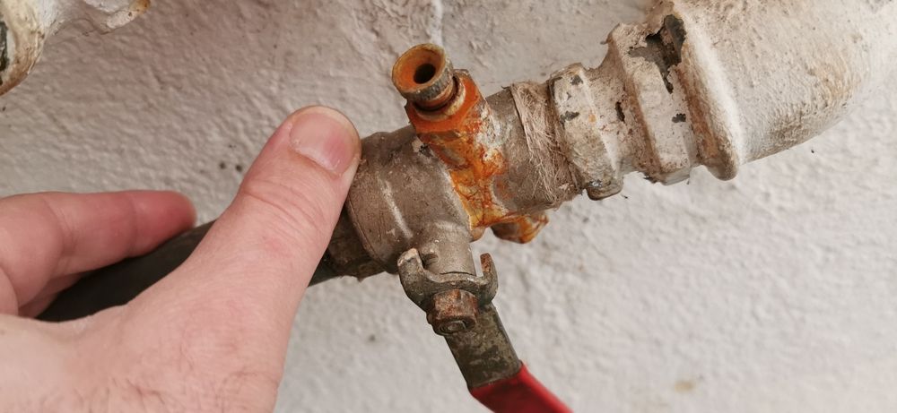 Hand holding a corroded pipe fitting. Rust is visible. Red valve handle. Pale wall in background.