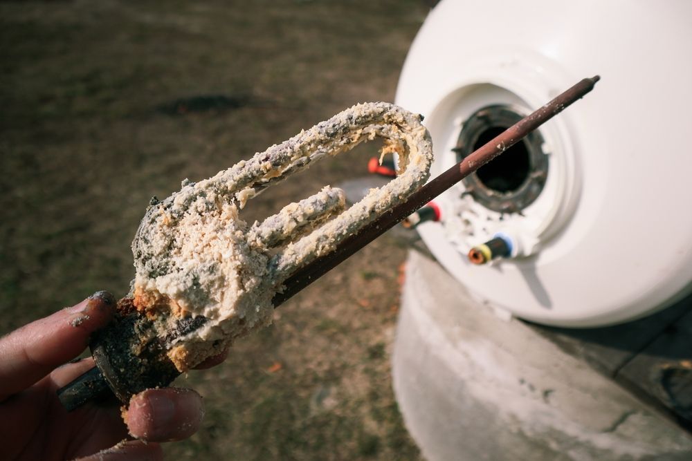 A heavily calcified water heater element being held, with the water heater tank visible in the background.