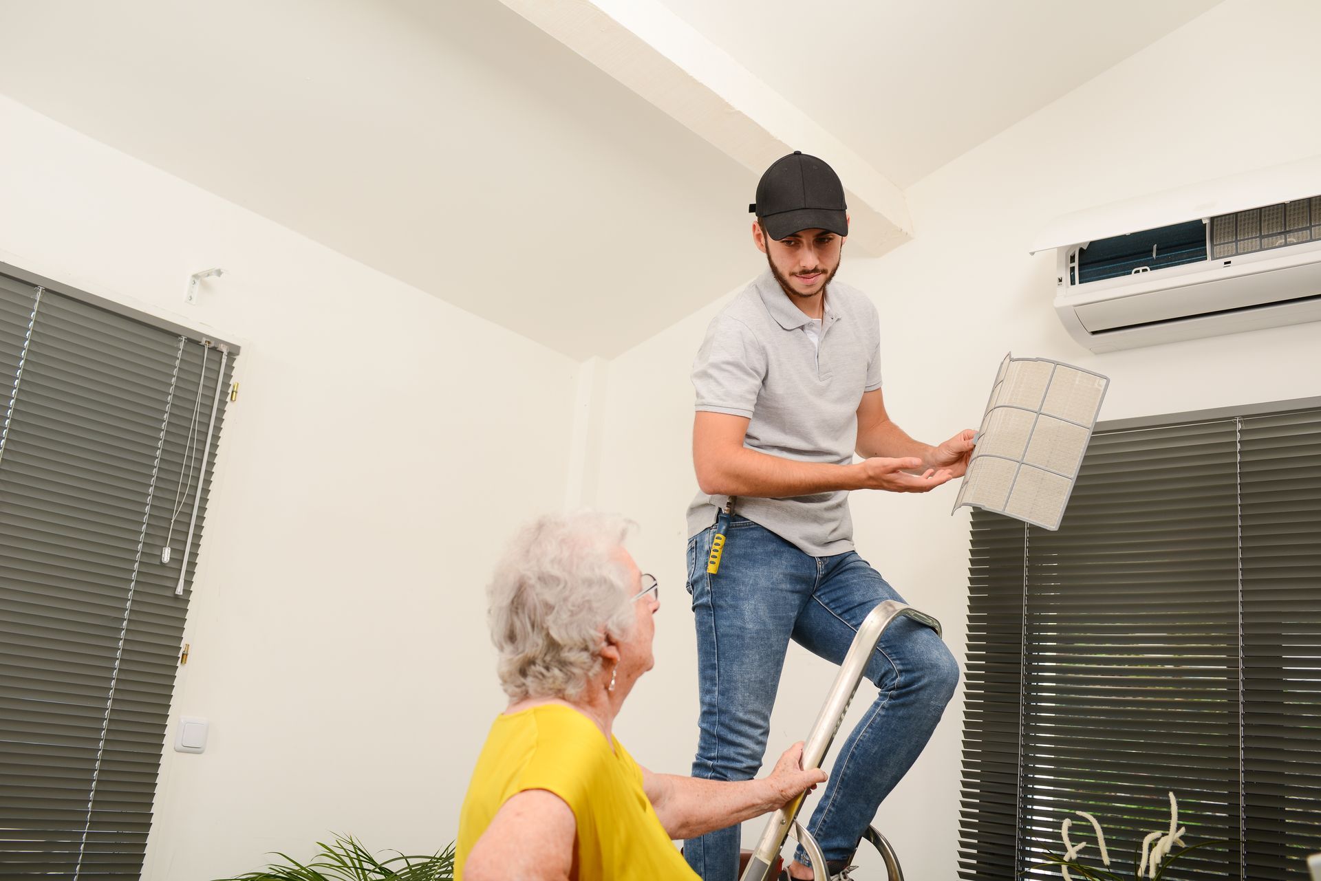 A man is standing on a ladder next to an older woman while fixing an air conditioner.