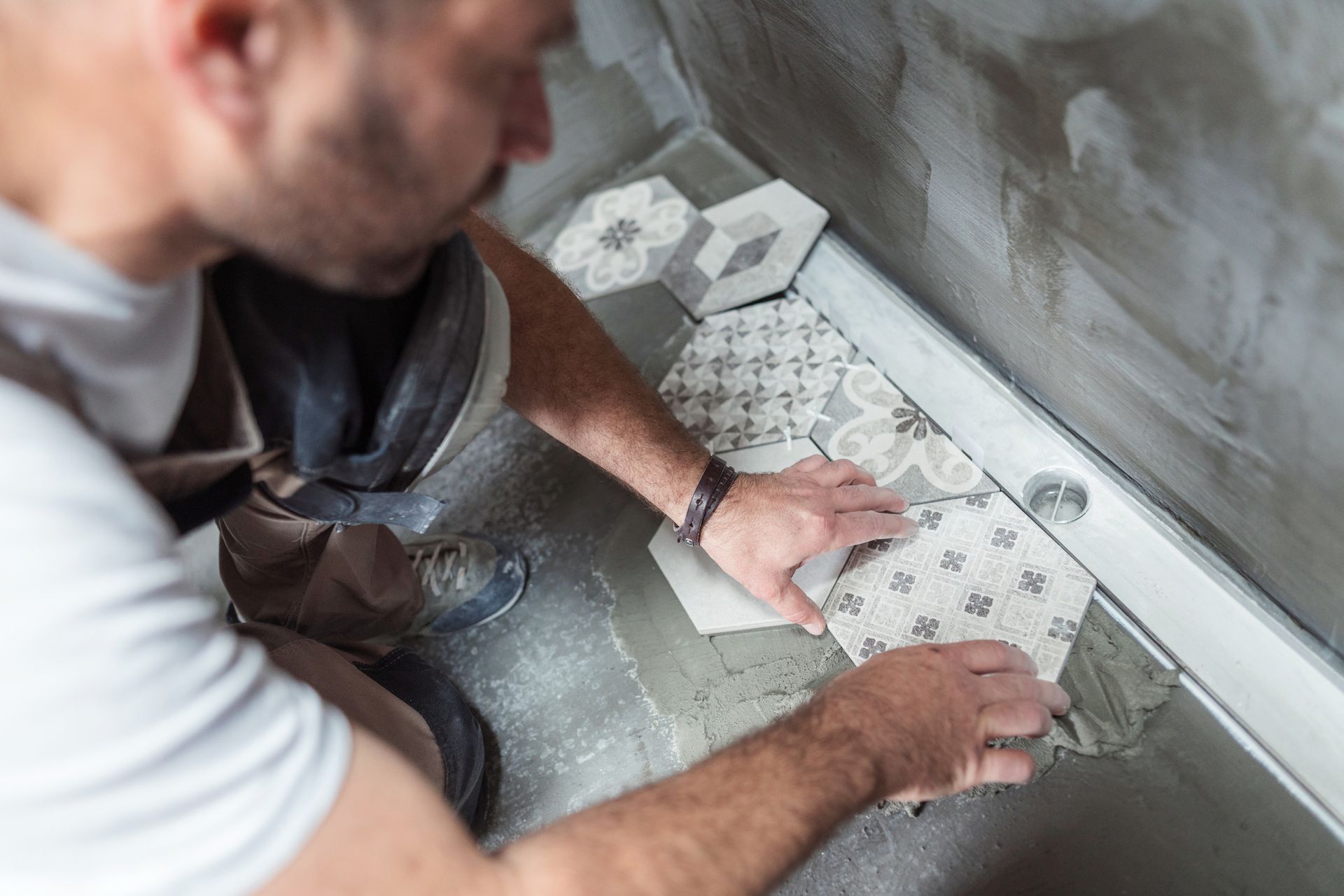 Man kneeling, installing patterned hexagon tiles in a shower. Gray cement, brown boots, light-colored tiles.
