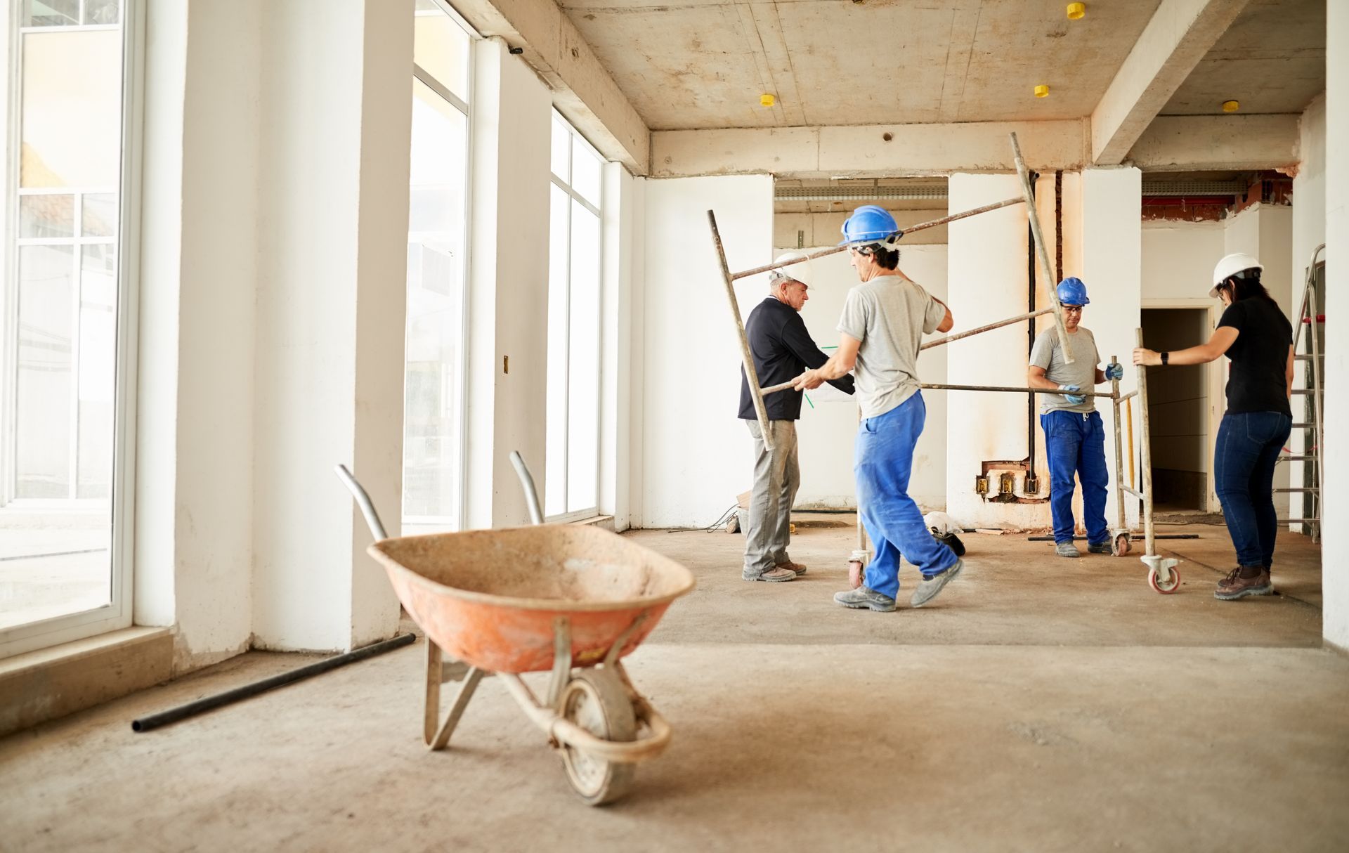 Construction workers inside a building, carrying poles, wearing hard hats. Wheelbarrow in foreground.