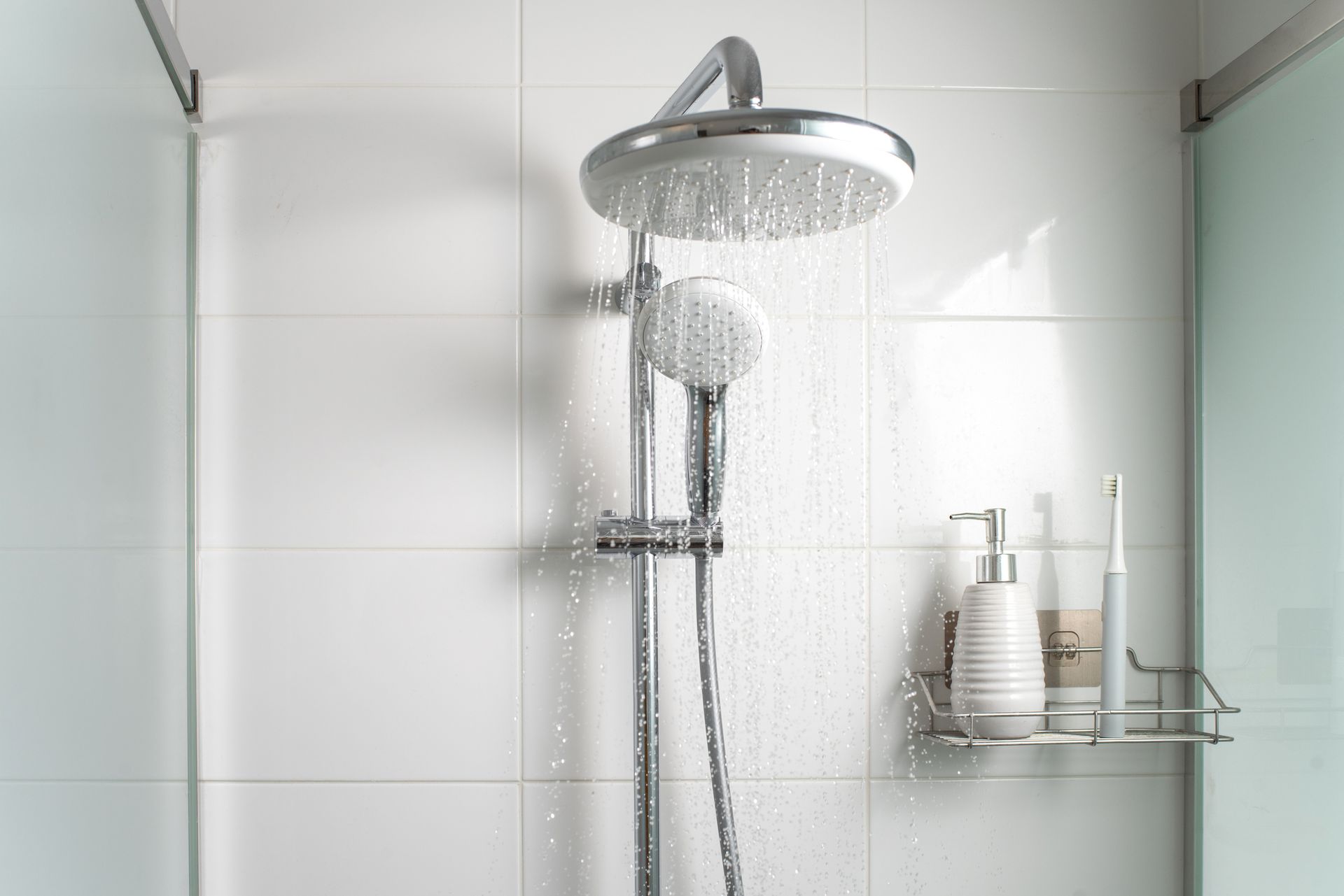 Shower head with water flowing, hand-held shower, shampoo and soap. White tiled wall.