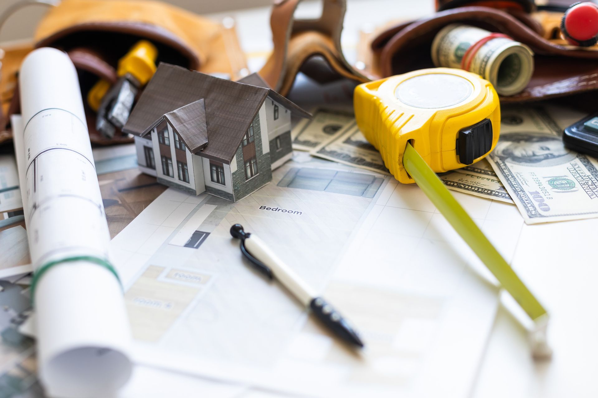 Construction tools and supplies on a desk: blueprints, house model, tape measure, money, and a tool belt.