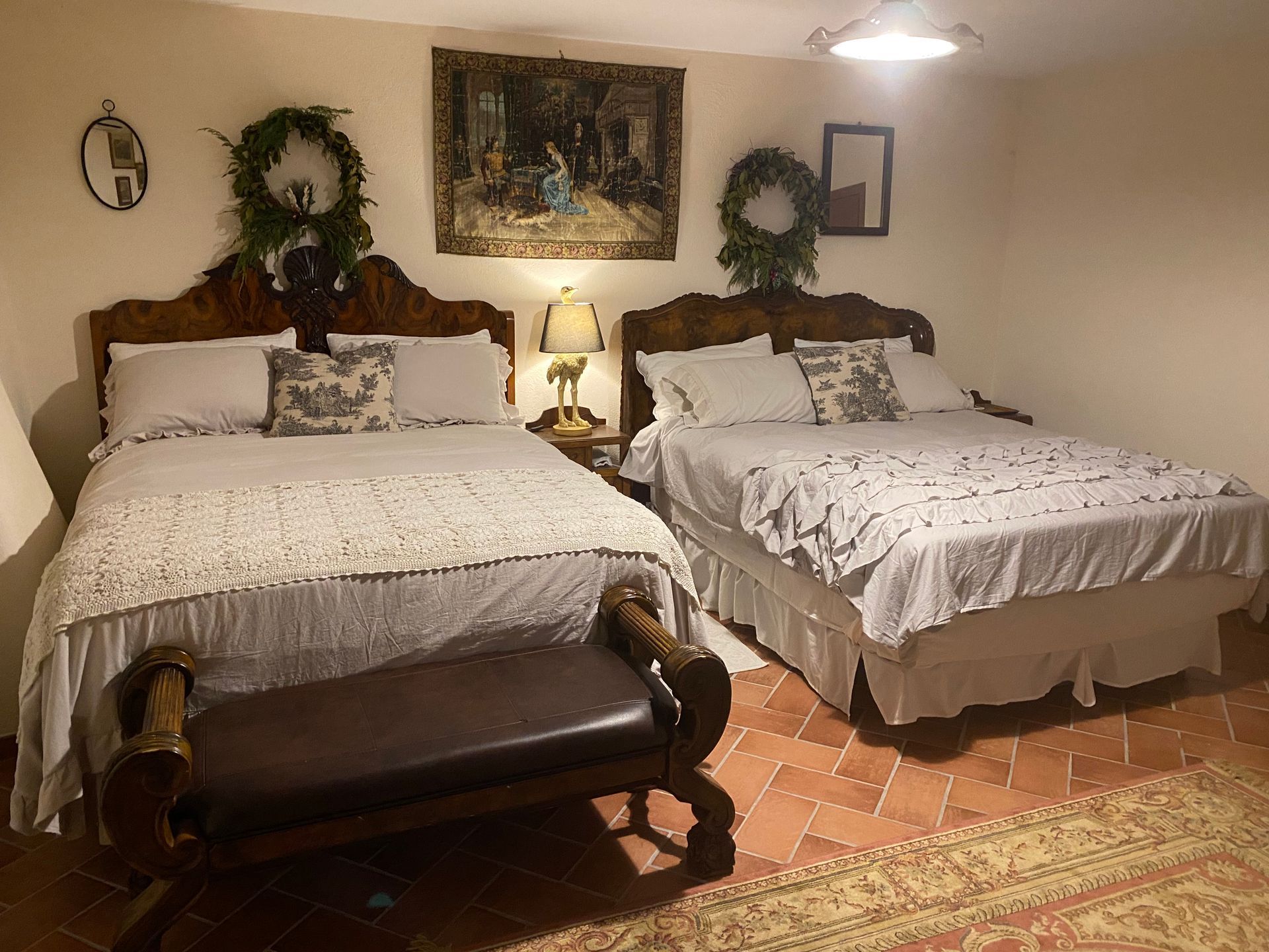 A bedroom with two queen beds, each with decorative wreaths and pillows, a central lamp, and a dark wood bench in front.