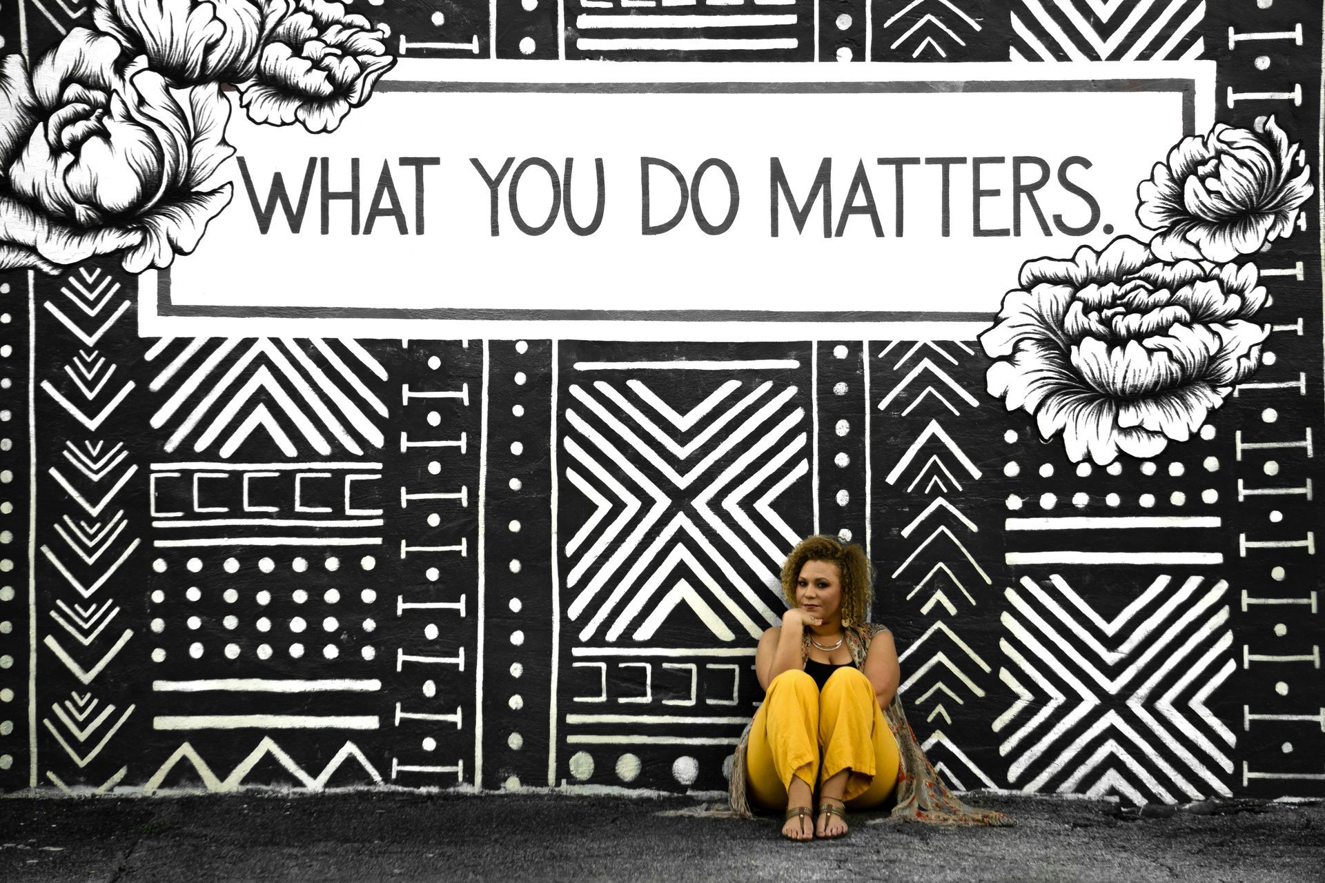 A woman in yellow pants sitting on the floor in front of a black-and-white tribal-decorated wall.