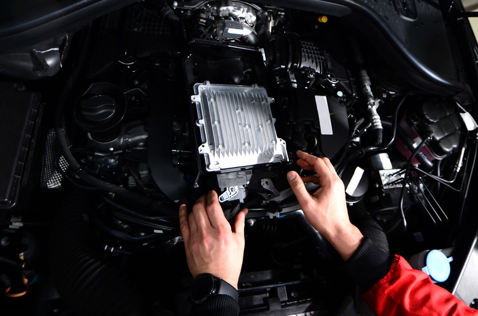 Mani che lavorano sul motore di un'auto, posizionando un componente rettangolare. Il motore è nero, illuminato dall'alto.