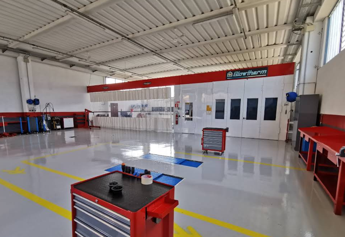 Ampia carrozzeria con dettagli rossi, cabina di verniciatura e vari utensili, illuminata da luci intense dall'alto.