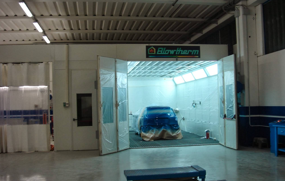 Auto in cabina di verniciatura, pronta per la verniciatura. La cabina ha porte pieghevoli aperte. Pareti e soffitto bianchi.