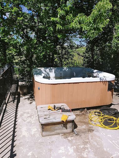 Hot tub on a concrete patio with tools and a yellow hose. Trees and a hillside are in the background.
