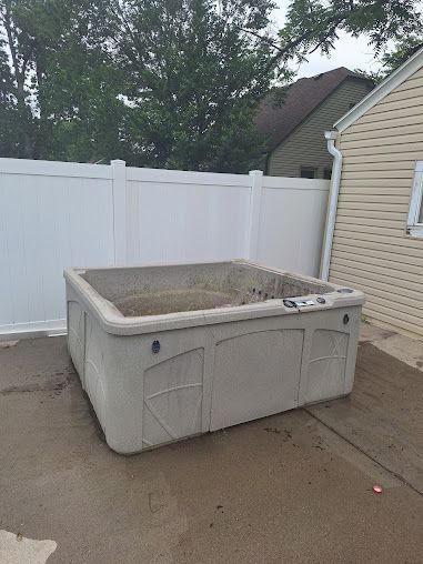 Square, tan hot tub on a concrete patio, next to a white fence and a beige house.