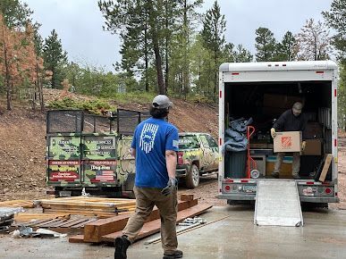 Two people loading boxes into a moving truck outdoors. Green dumpsters and a vehicle are in the background.