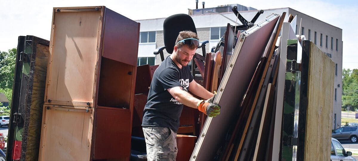 A person loads discarded wood and furniture into a truck. Sunlight shines on a building in the background.
