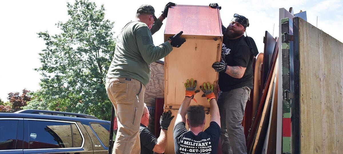 Four people lifting a large wooden box into a truck bed. Outdoors, with a tree and a vehicle visible.