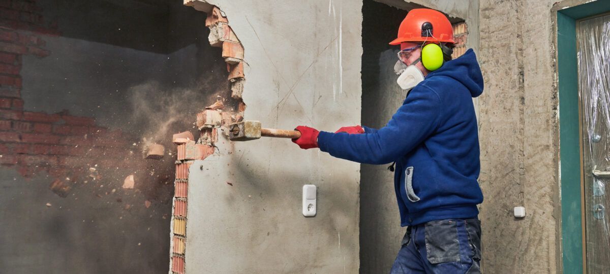 Person in safety gear demolishes a brick wall with a sledgehammer, creating a hole in a building.