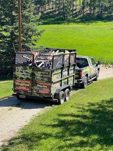 Camouflage truck and trailer loaded with debris on a gravel road, green grass and trees in background.