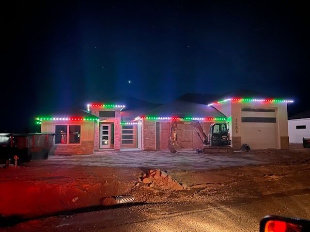House with red, green, and white Christmas lights along the roofline at night; construction equipment in front.