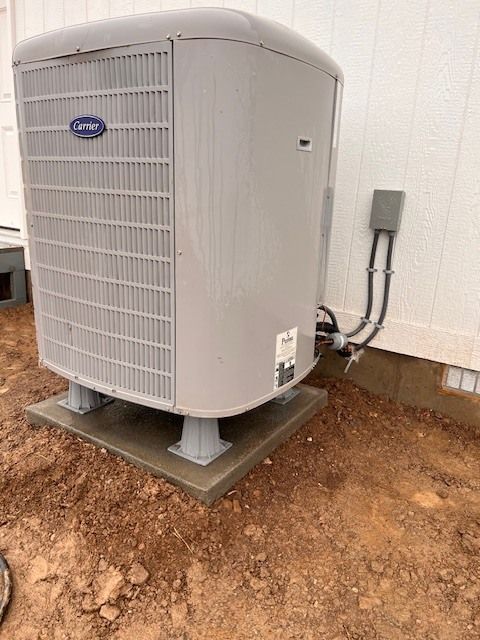 An air conditioning unit on a concrete pad with dirt ground. Gray unit with electrical conduit on side.