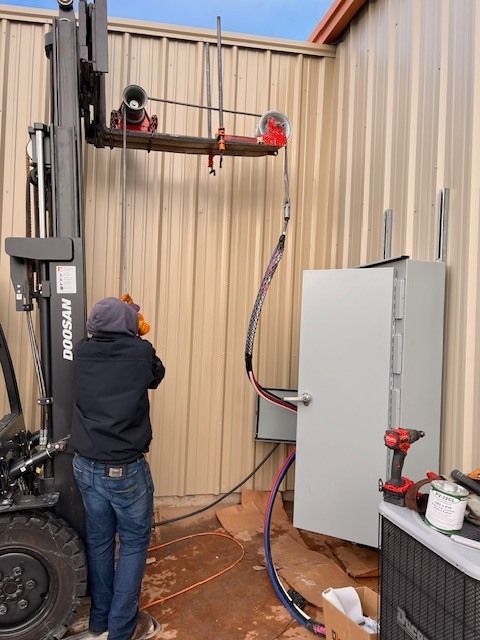 Man working on electrical box with forklift assisting, outdoors.