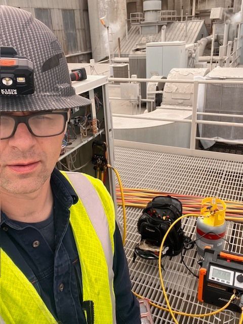 Person in hard hat and safety vest on industrial site, taking selfie. Electrical equipment visible.