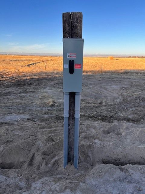 Electrical box on a wooden post in an open, sandy field under a blue sky.