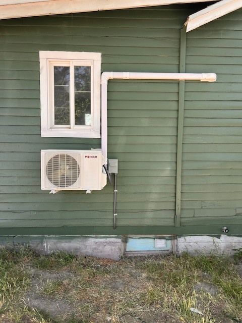 White air conditioner unit mounted on green building exterior, near a window. White pipe extends from unit.