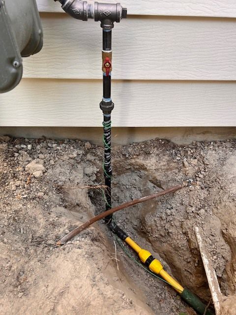 A gas pipe exposed in a trench next to a beige house, with a section wrapped in green tape.