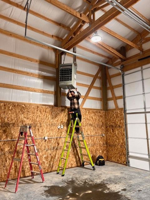 Person on a ladder installing a heater in a garage. Wood walls, light fixture, and metal conduit are visible.