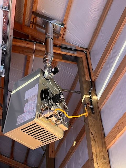 A gray, suspended gas heater with a chimney in a wooden building, connected to a gas line.
