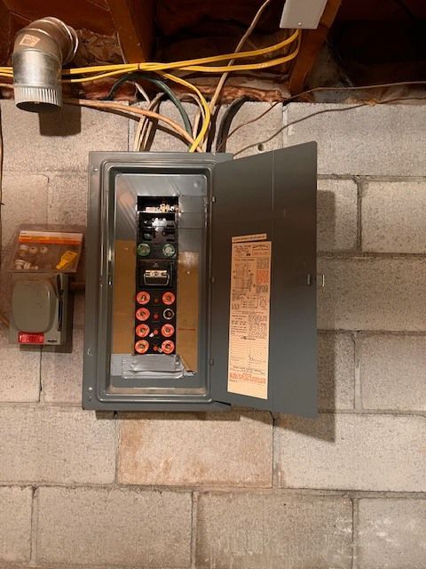 Gray electrical panel with open door mounted on a concrete block wall. Wires and conduit are visible.
