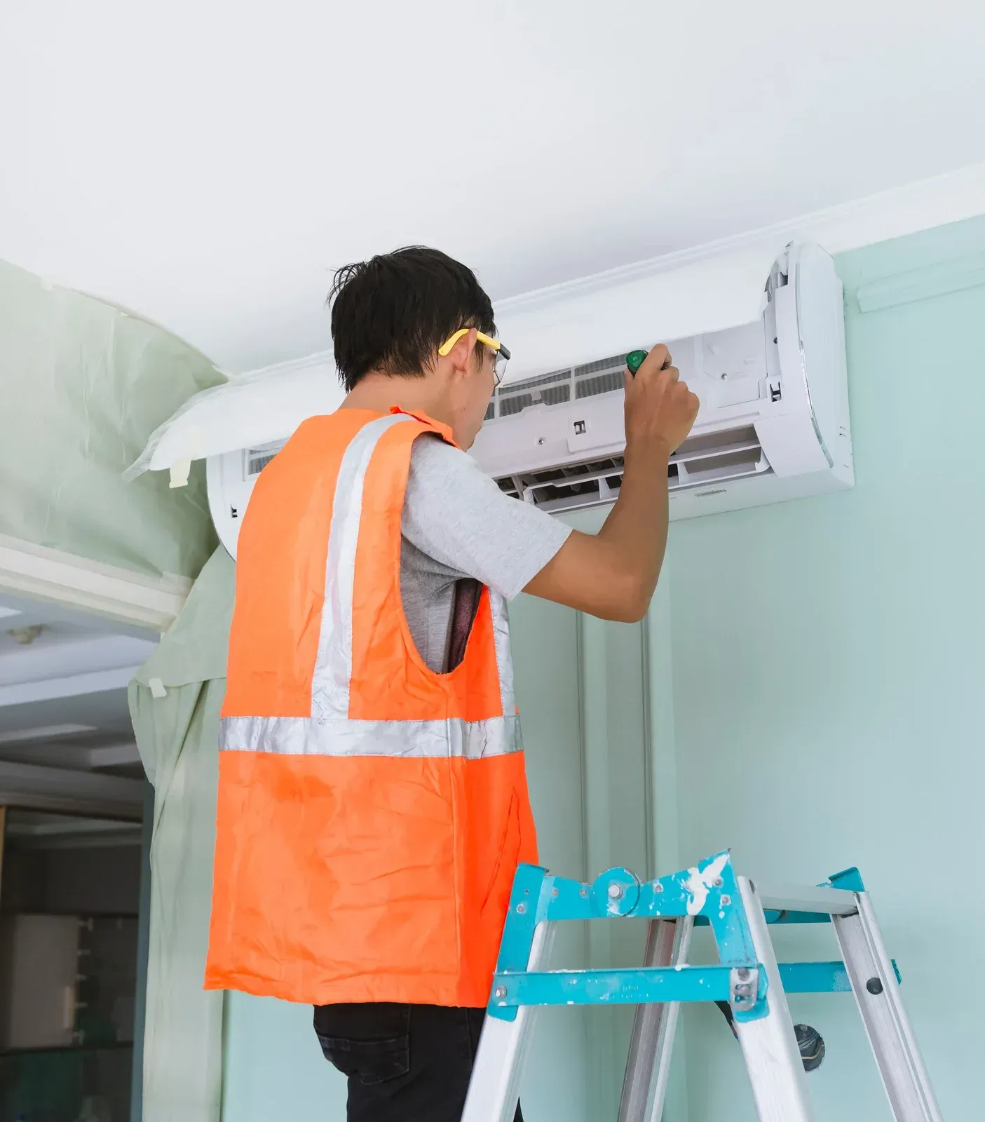 HVAC technician in orange vest on a ladder, repairing a white wall-mounted air conditioner.