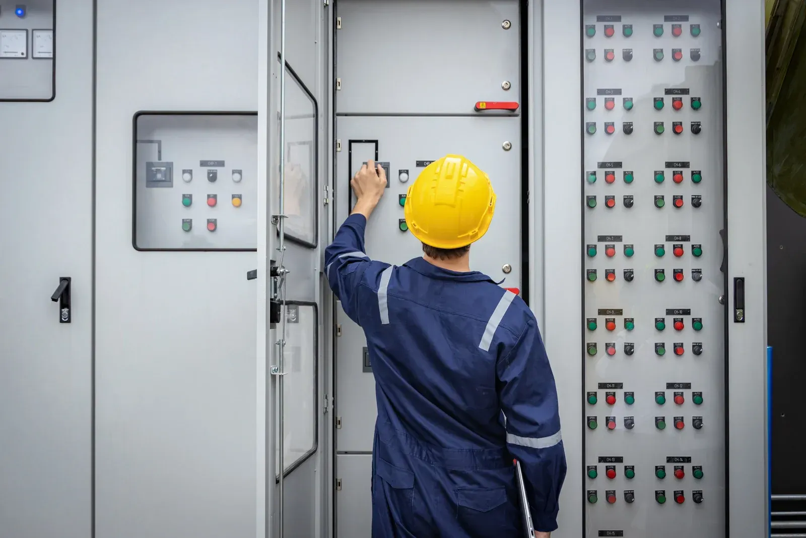 Electrician in blue coveralls and yellow helmet, inspecting electrical panel.