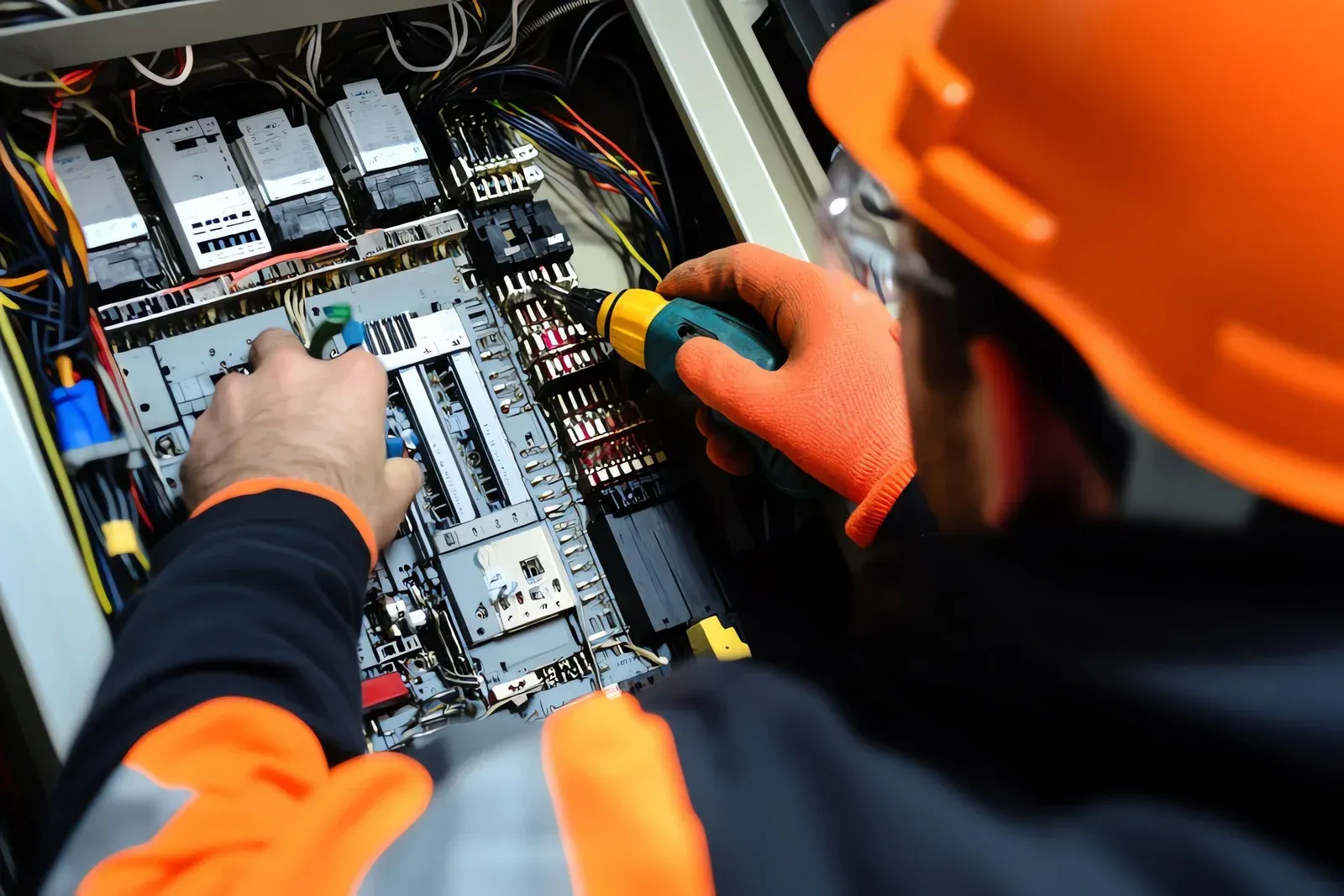 Electrician in orange safety gear, inspecting electrical panel wiring with a tool.