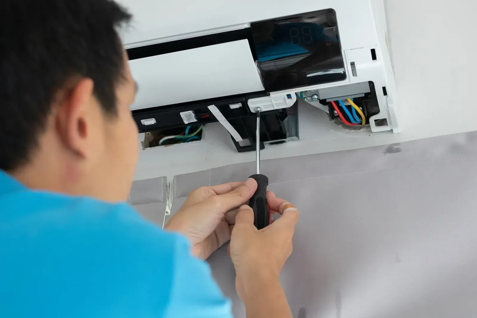 Person using a screwdriver to repair a white air conditioning unit mounted on a wall.