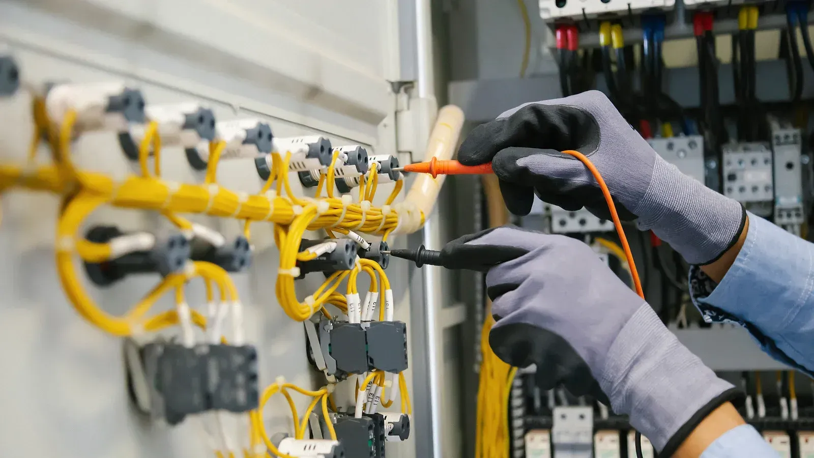 Electrician in gloves testing wires in a control panel with a multimeter.