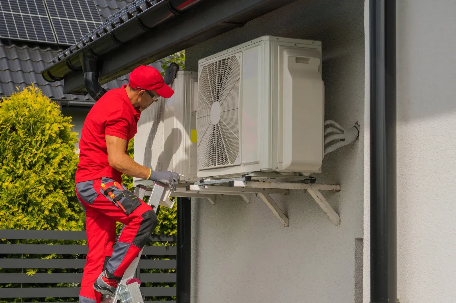 A worker in red installs an outdoor air conditioning unit on a white wall.