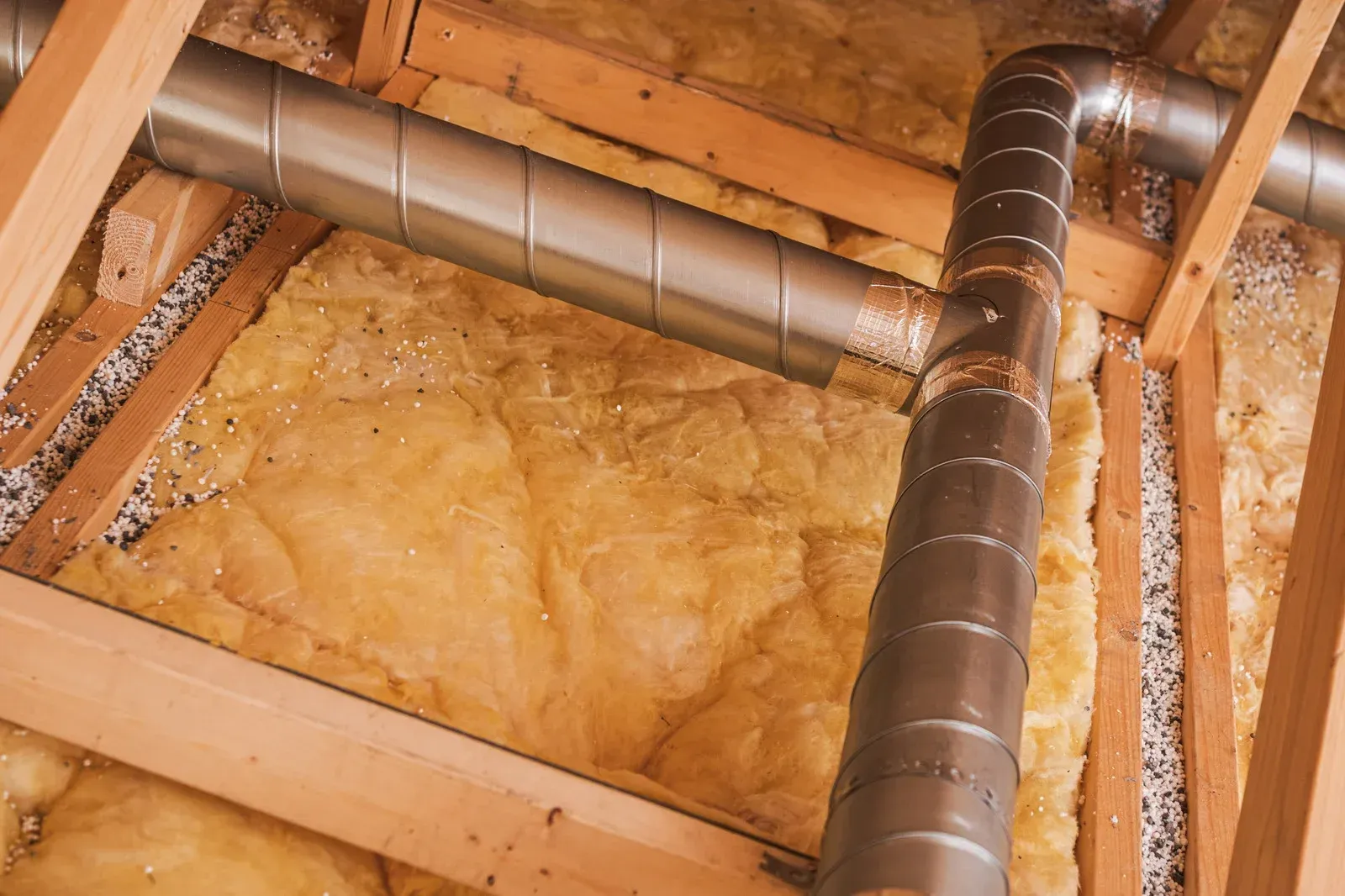 Wooden attic framing with insulation, a silver duct, and other ducting.