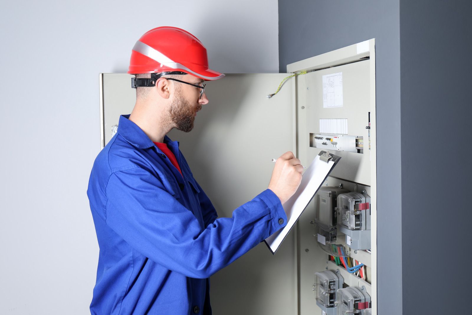 Electrician in a blue jumpsuit and red hard hat inspecting an electrical panel, writing on a clipboard.