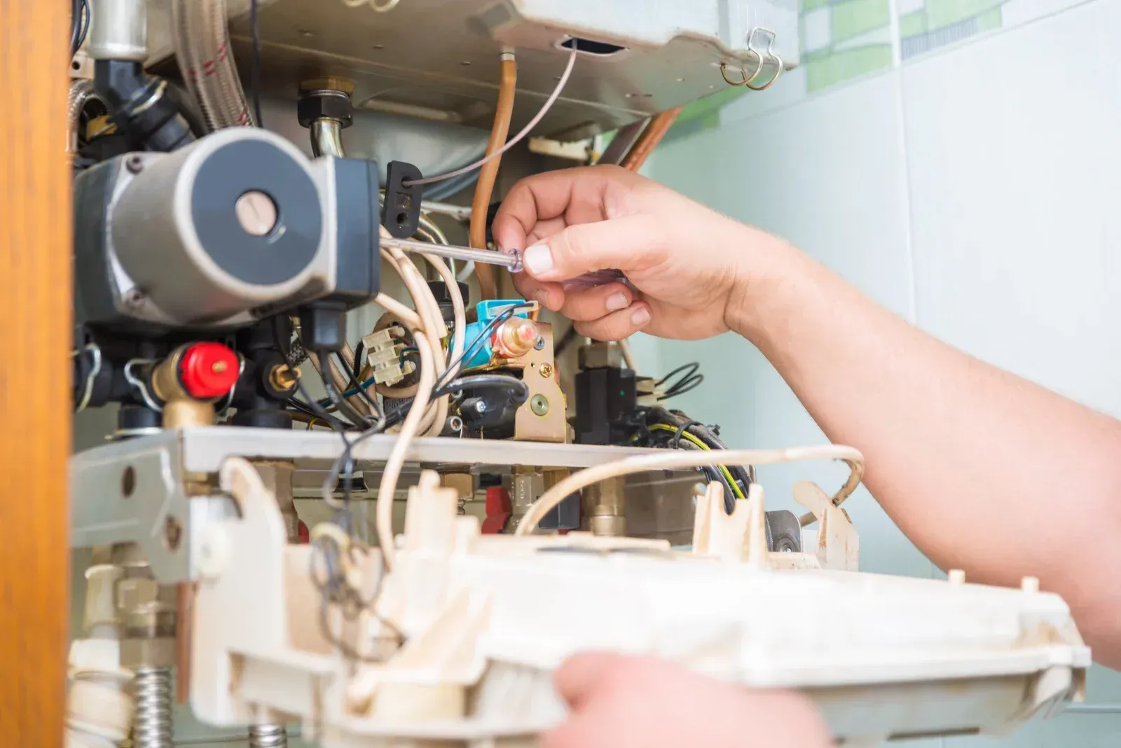 Person repairs a white boiler, using a screwdriver, in a tiled room.