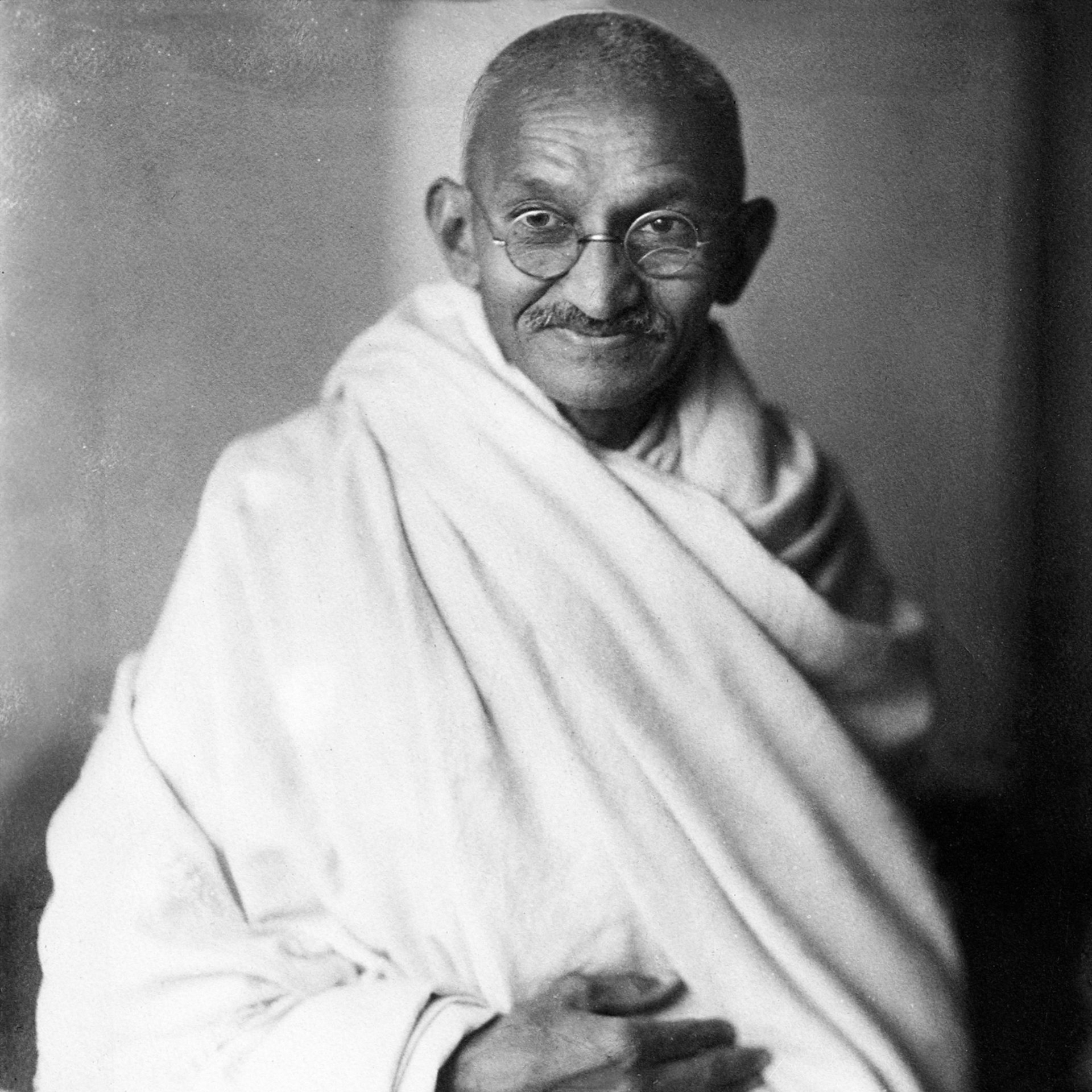 Mahatma Gandhi, met een ronde bril en een sjaal om, lacht naar de camera.