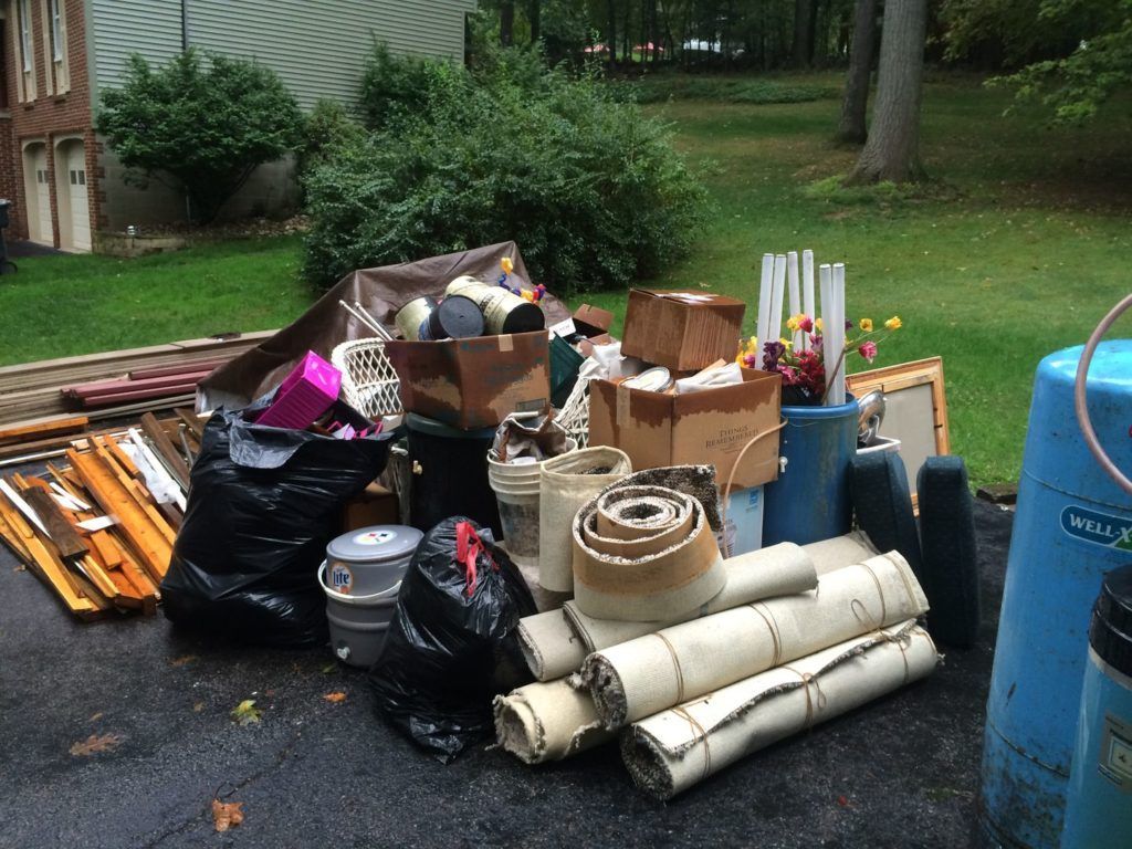 A pile of household junk, including black trash bags, cardboard boxes, and rolled carpets, on a driveway near grass.