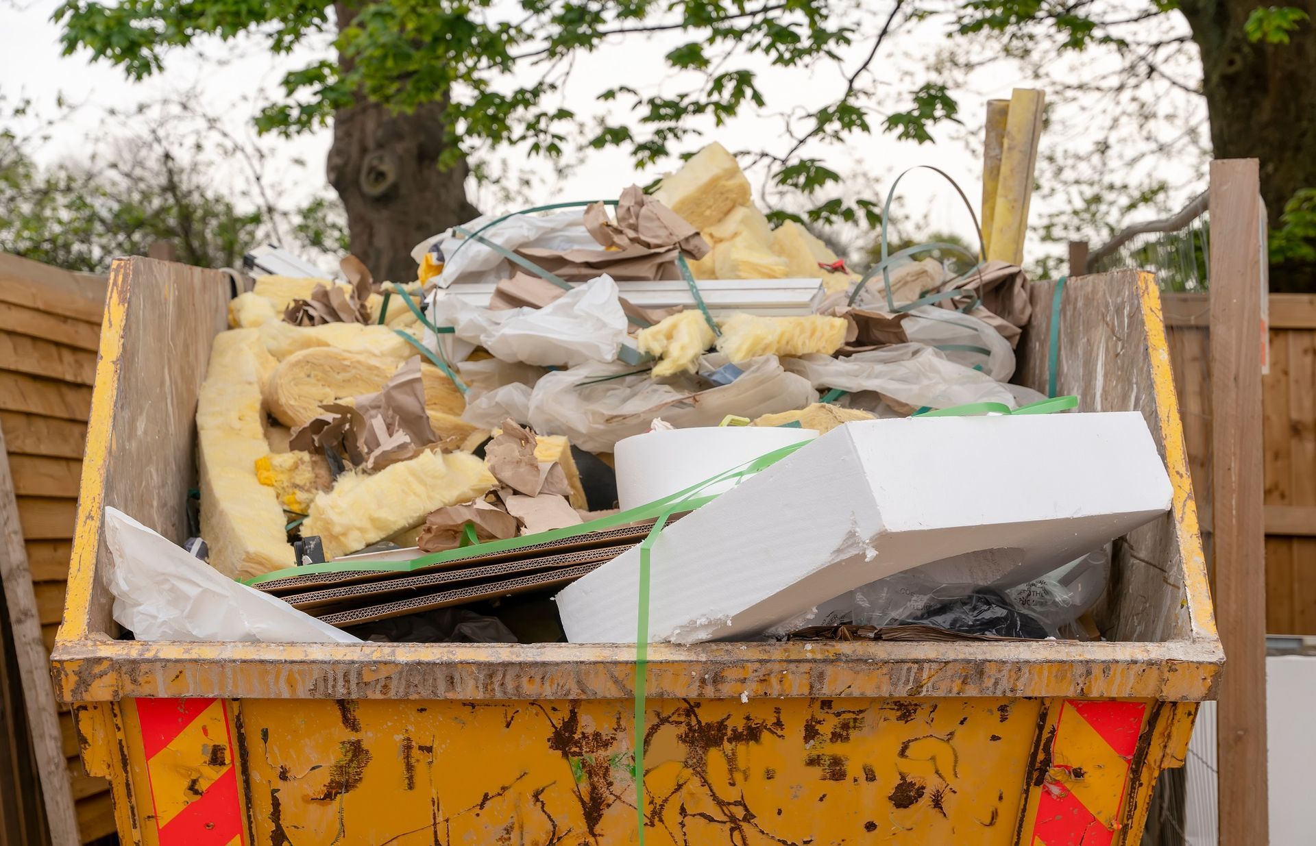 A yellow construction skip filled with various construction debris, foam, and cardboard, set against a backdrop of trees.